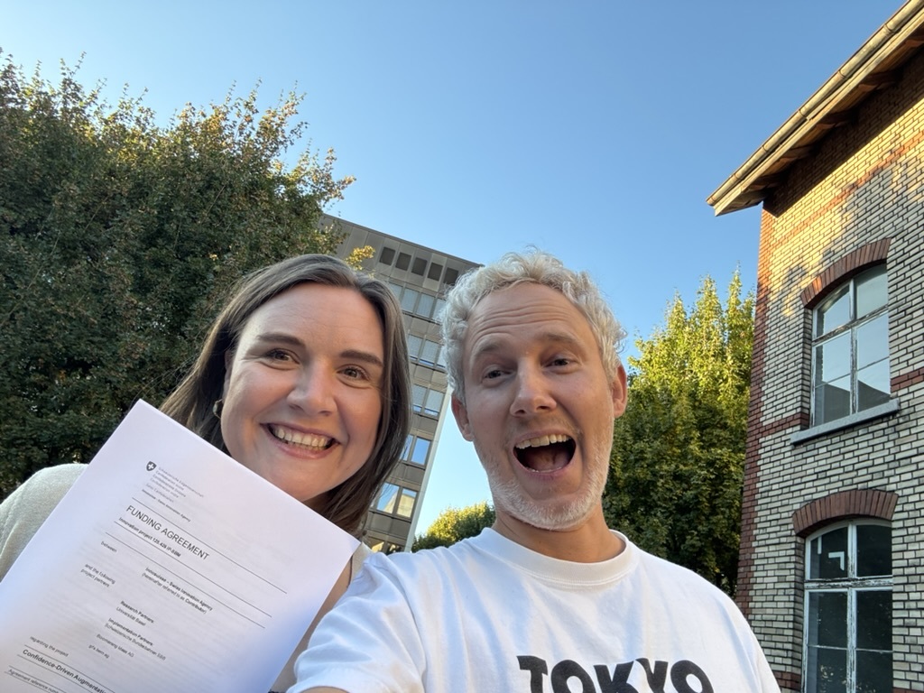 Two people taking a selfie outdoors, one holding up a printed letter